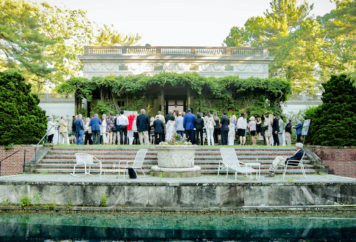 a group of people stand with their backs to the camera, in front of a stone building entrance framed by vines